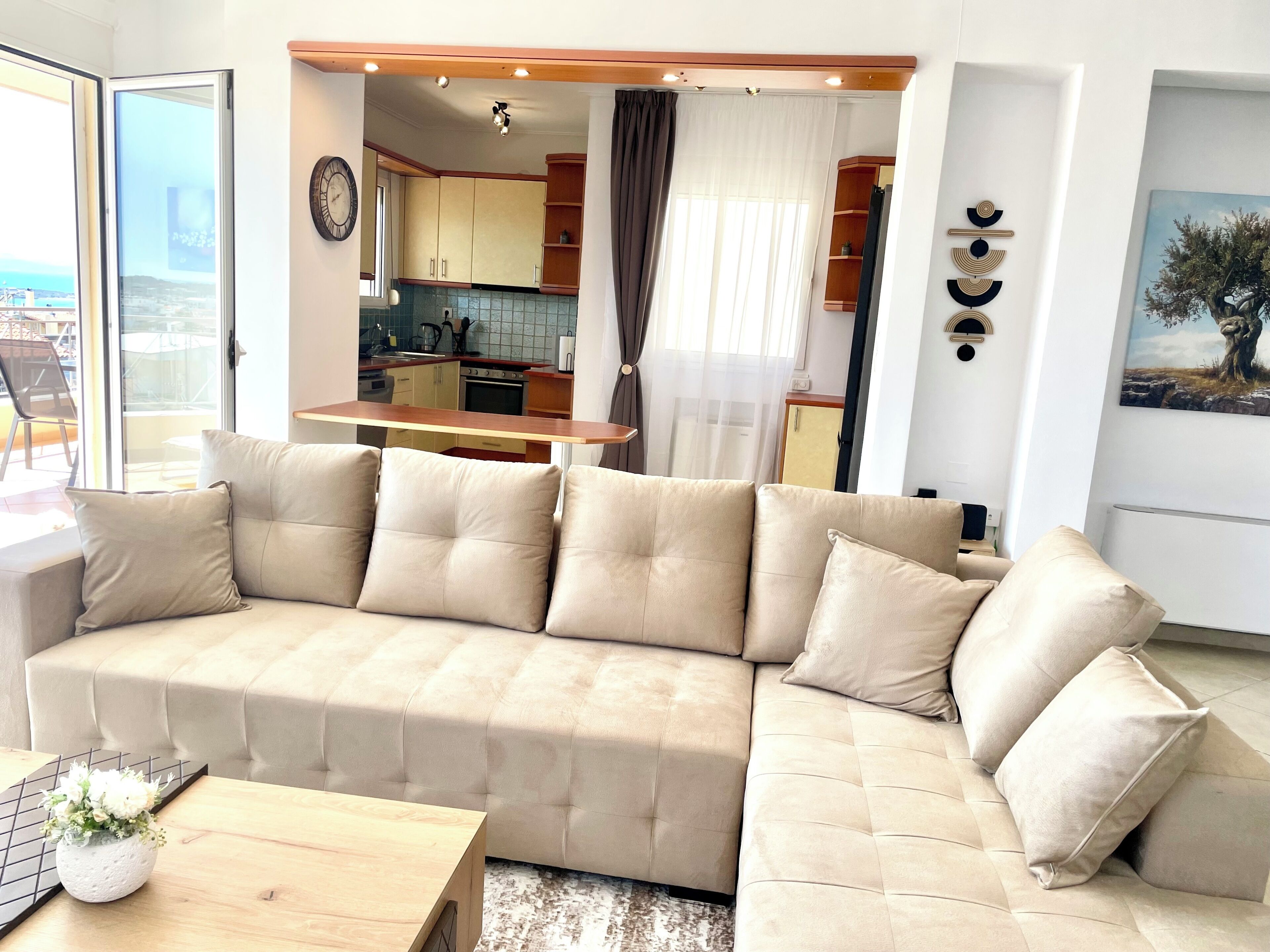 Un salon moderne avec un canapé d’angle beige, une table basse en bois et une petite plante en pot. On aperçoit une cuisine à l’arrière-plan, avec des meubles de cuisine en bois et une fenêtre.