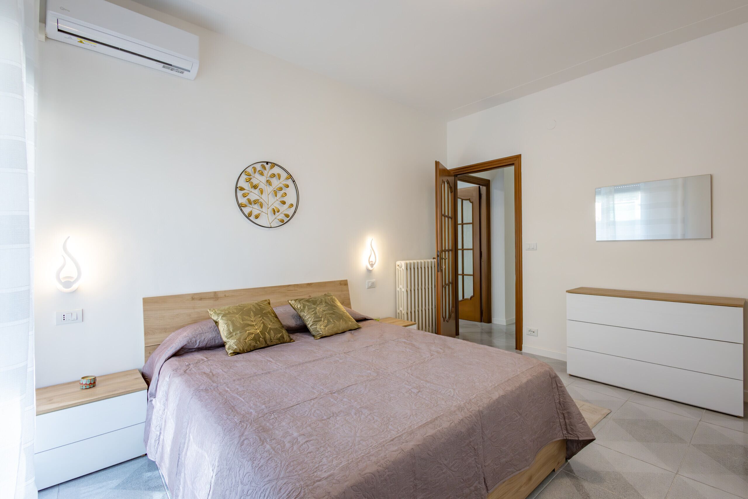 A bedroom with a bed, bedside tables, a dresser, a mirror, and an air conditioning unit.