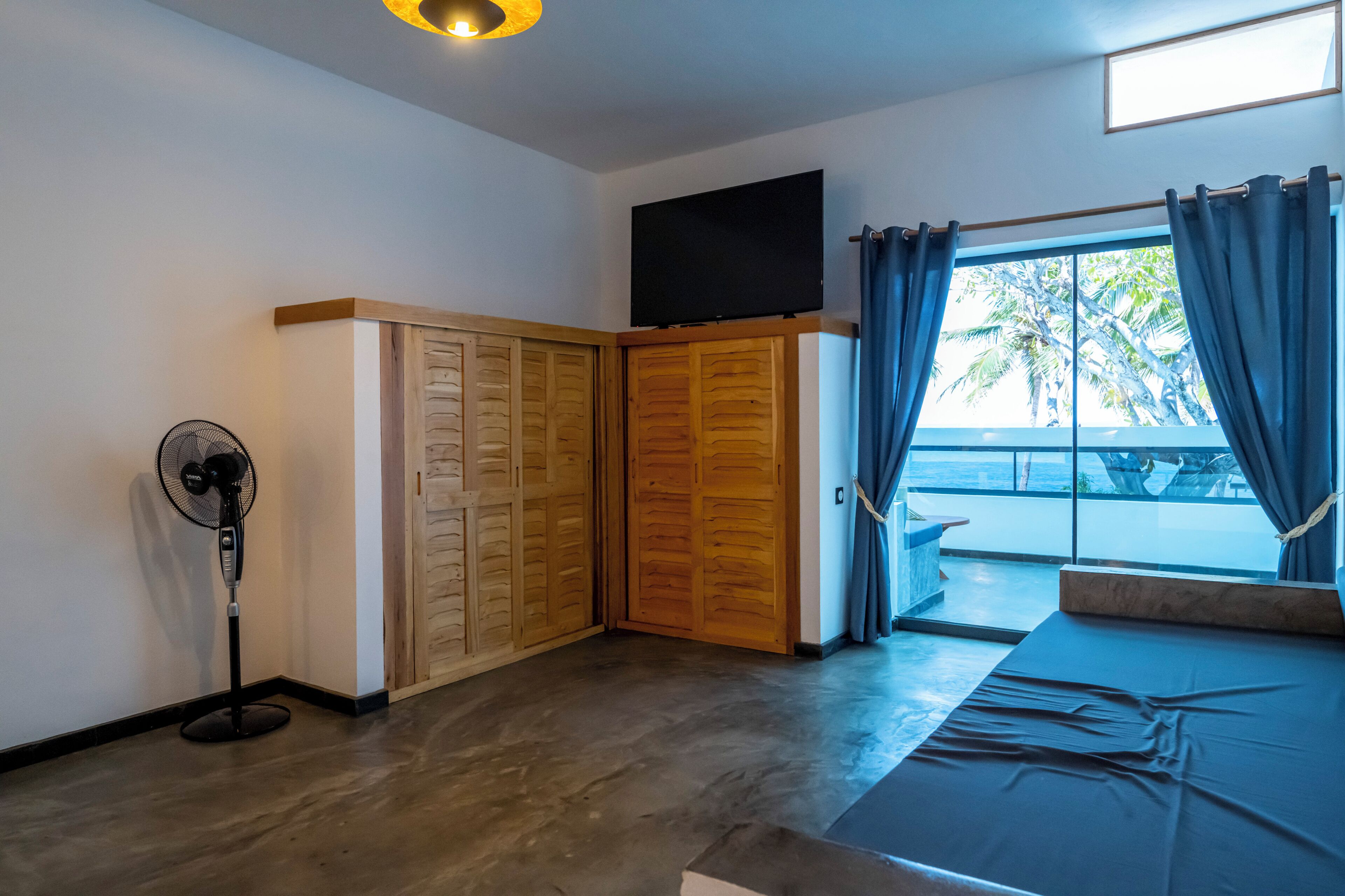 Una habitación con una cama, un ventilador, un televisor y vistas a palmeras a través de una ventana.