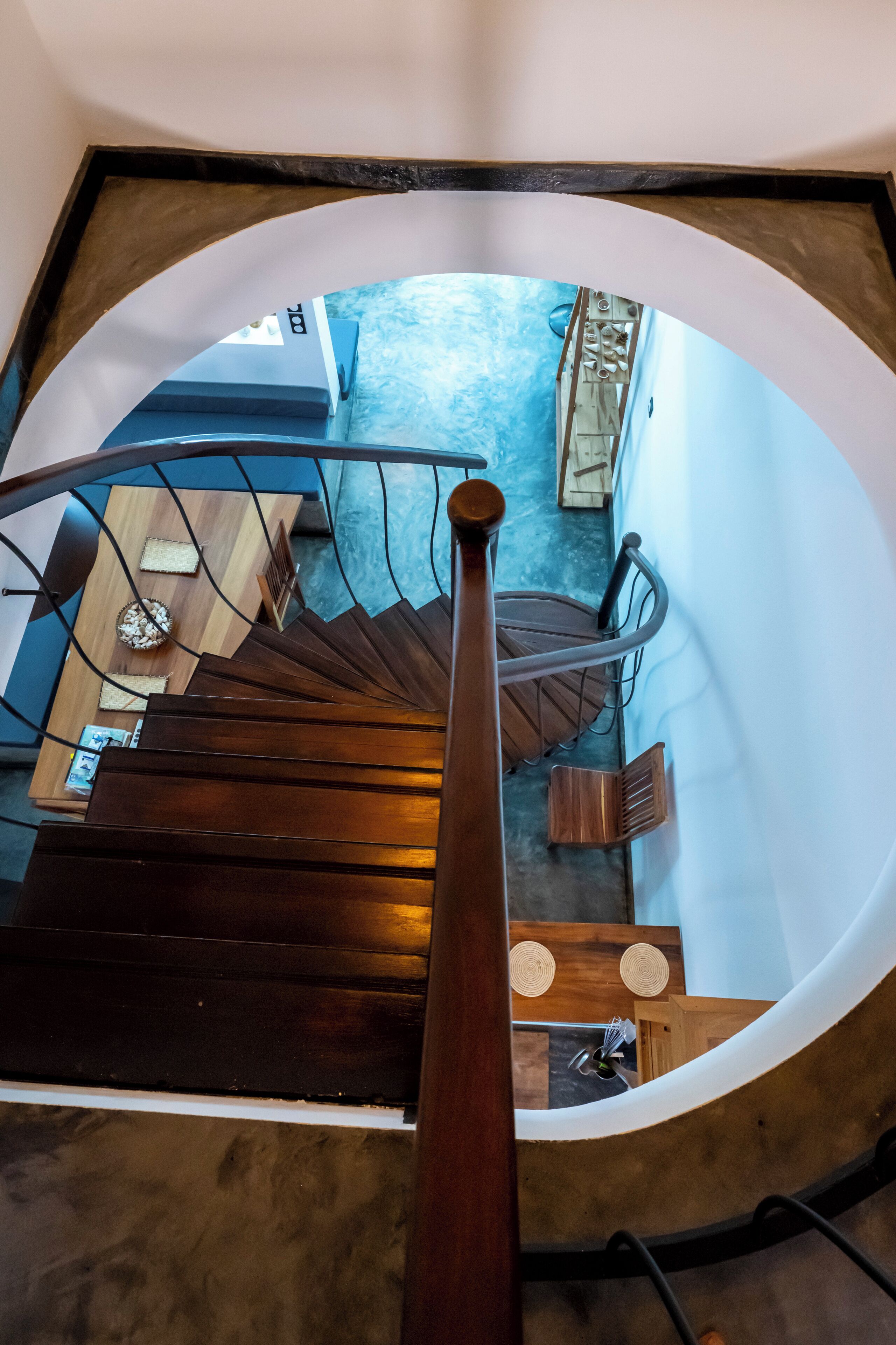 A spiral staircase with a wooden handrail, leading to a room with a blue textured floor and a wooden table.