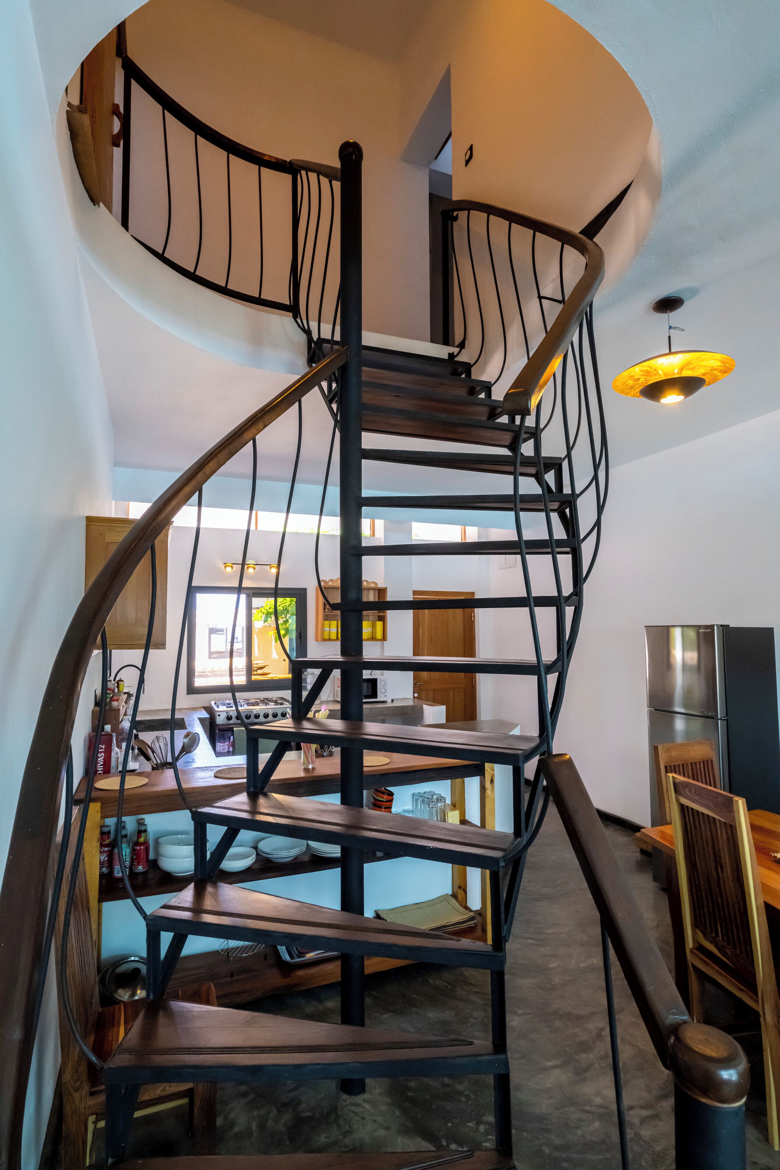 Una escalera de caracol con barandilla de metal que conduce a un área del segundo piso con cocina y comedor.