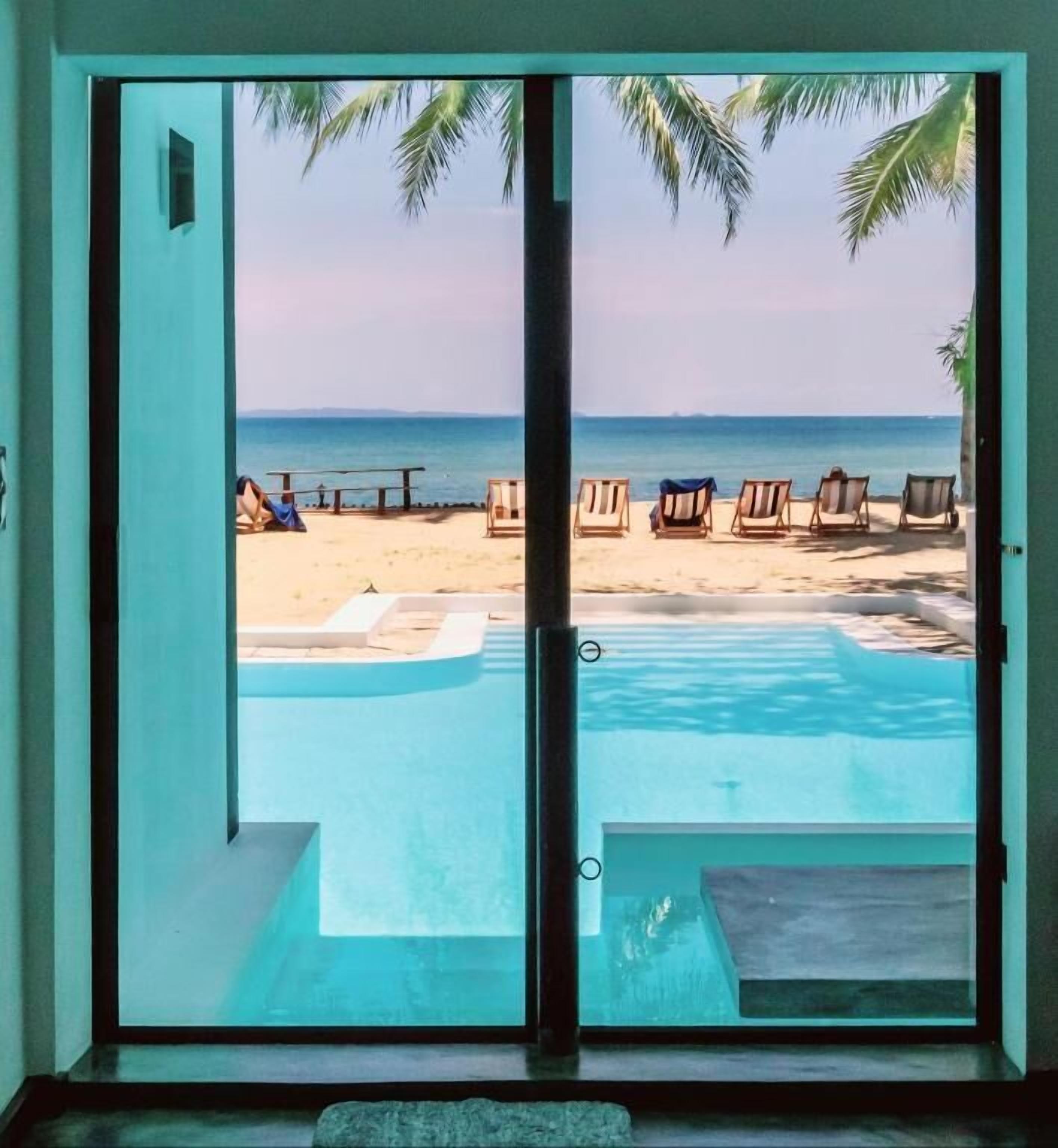A view through a sliding door of a swimming pool with lounge chairs and a beach in the background.