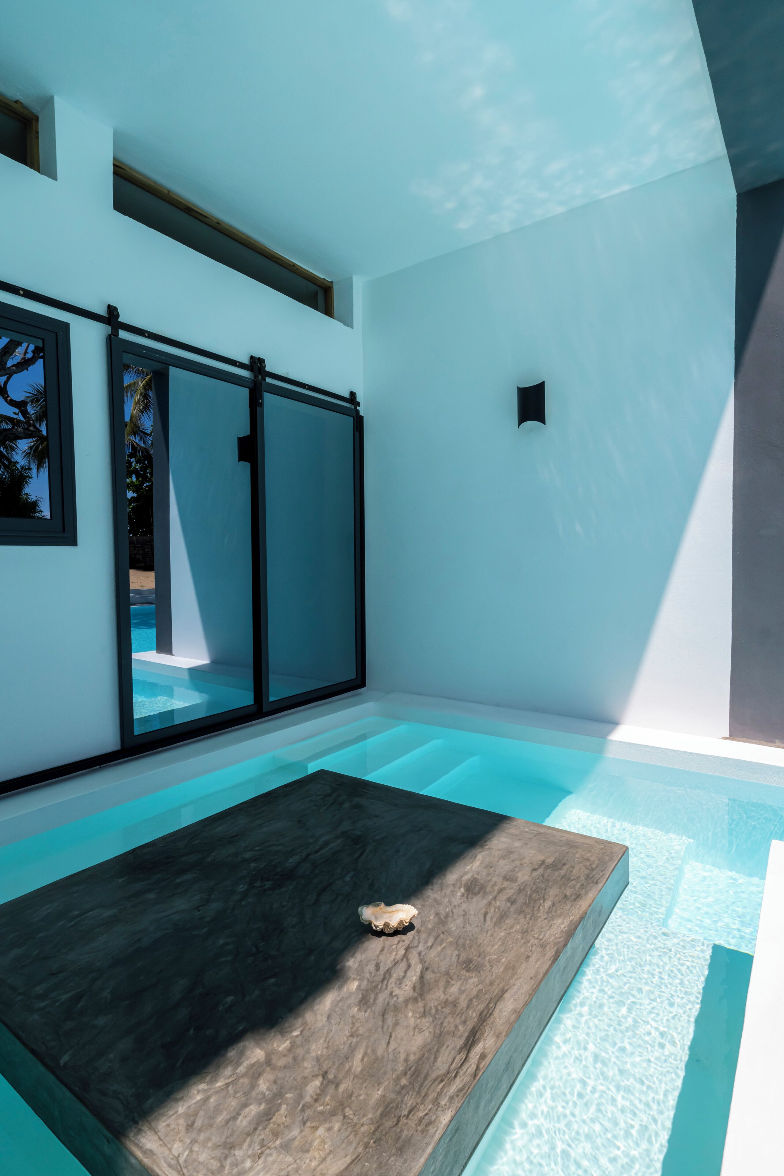 Una piscina interior moderna con agua cristalina, una plataforma de madera y grandes puertas correderas de vidrio.