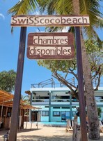 Letrero que indica 'SwissScocobeach' y 'chambres disponibles', con vista a un edificio moderno y palmeras.