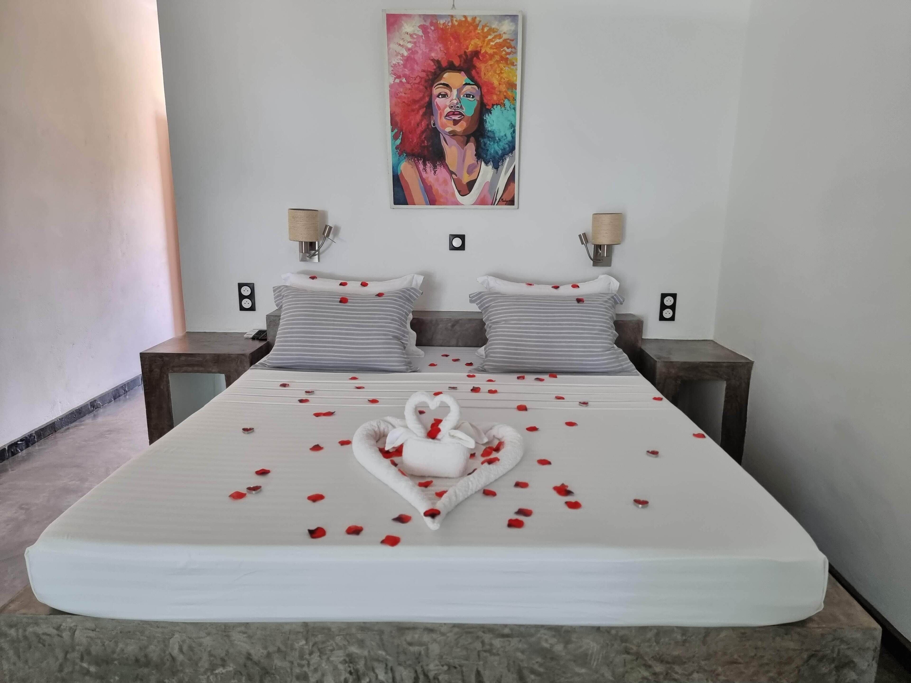 Une chambre d’hôtel avec un grand lit, deux tables de chevet, un tableau accroché au mur et un cygne décoratif entouré de pétales de rose.