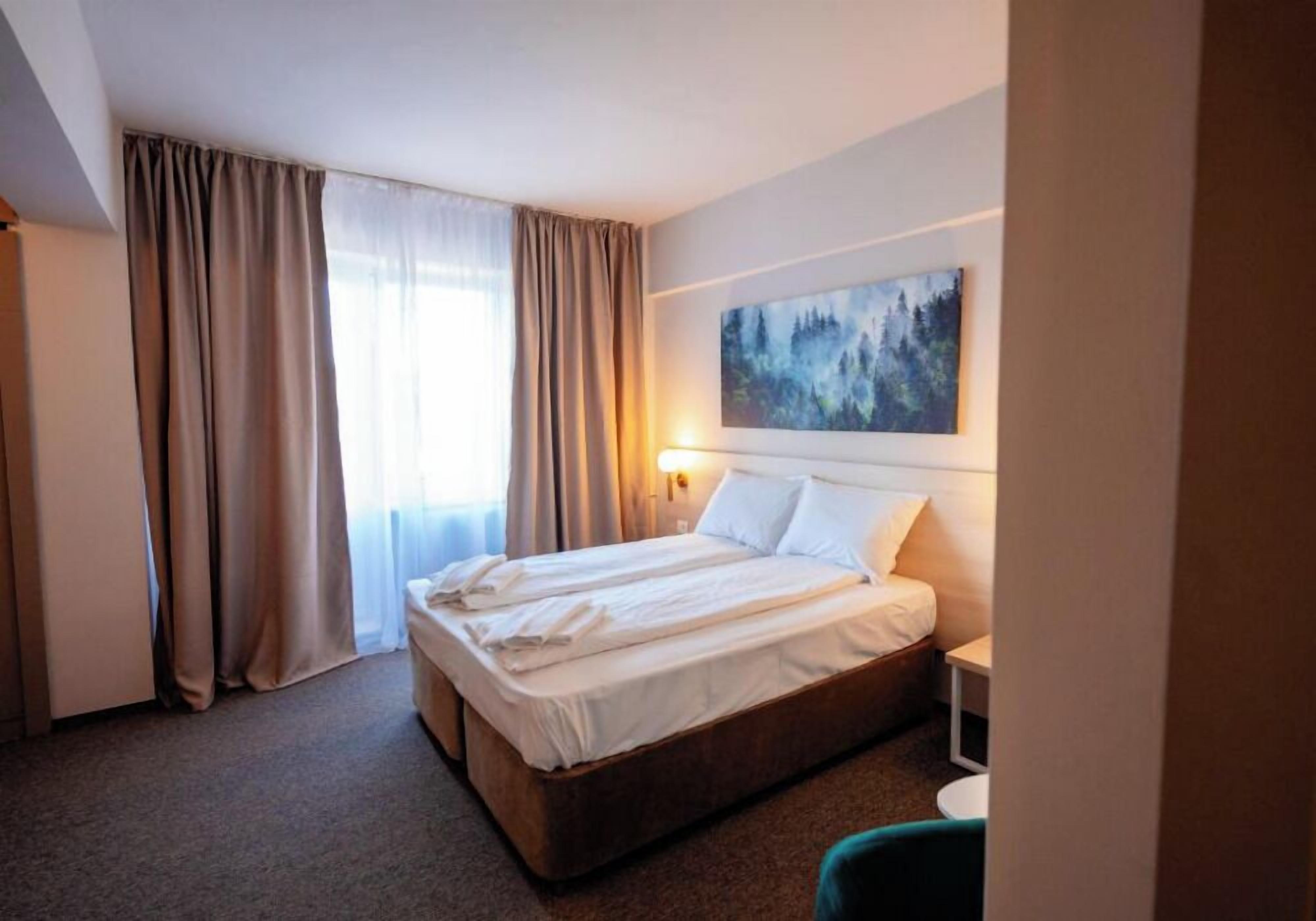 A hotel room with a bed, curtains, a painting on the wall, and a nightstand.