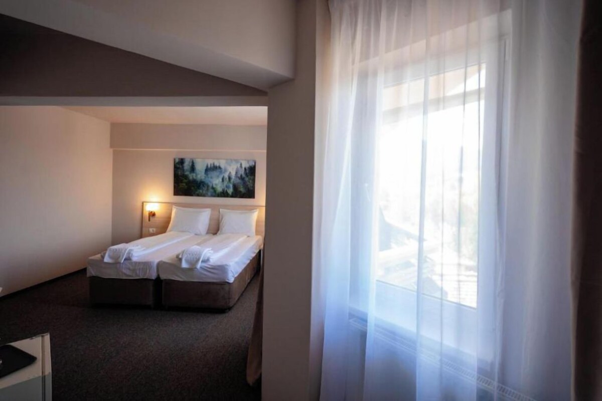 A hotel room with a bed, a desk, a painting on the wall, and a window with sheer curtains.