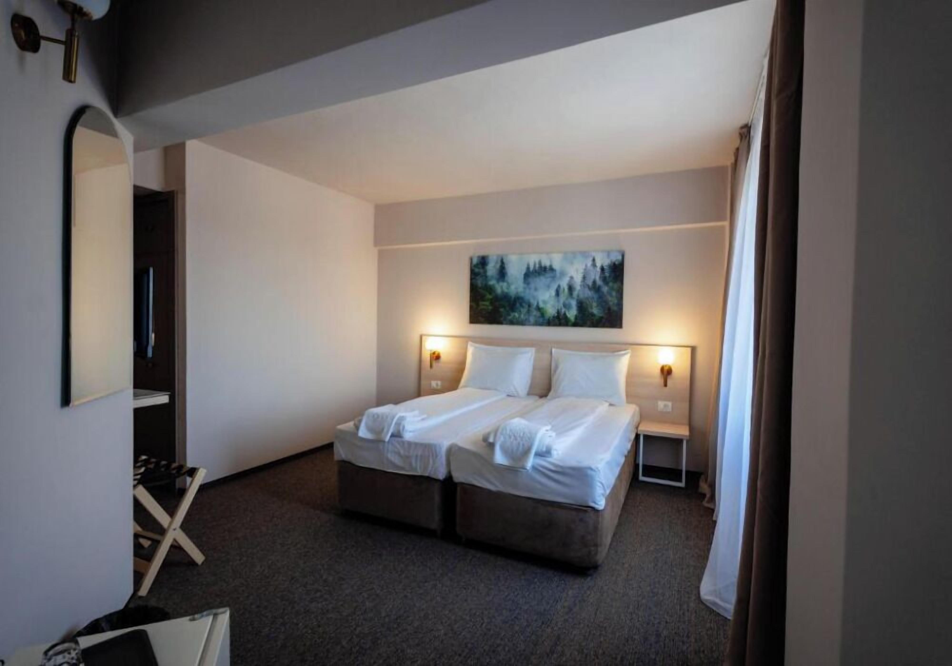 A hotel room with a double bed, a painting on the wall, and a nightstand with a lamp.