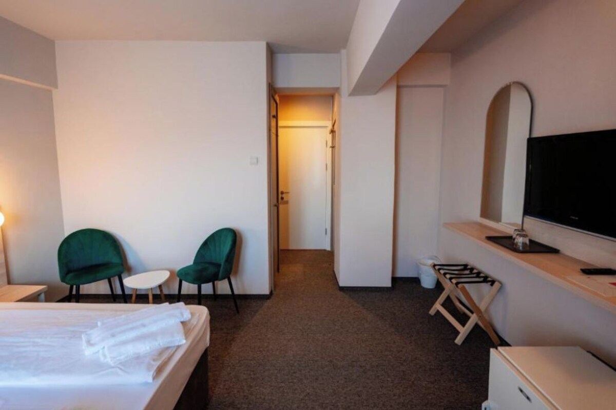 A hotel room with a bed, two green chairs, a small table, a TV, and a desk with a computer.