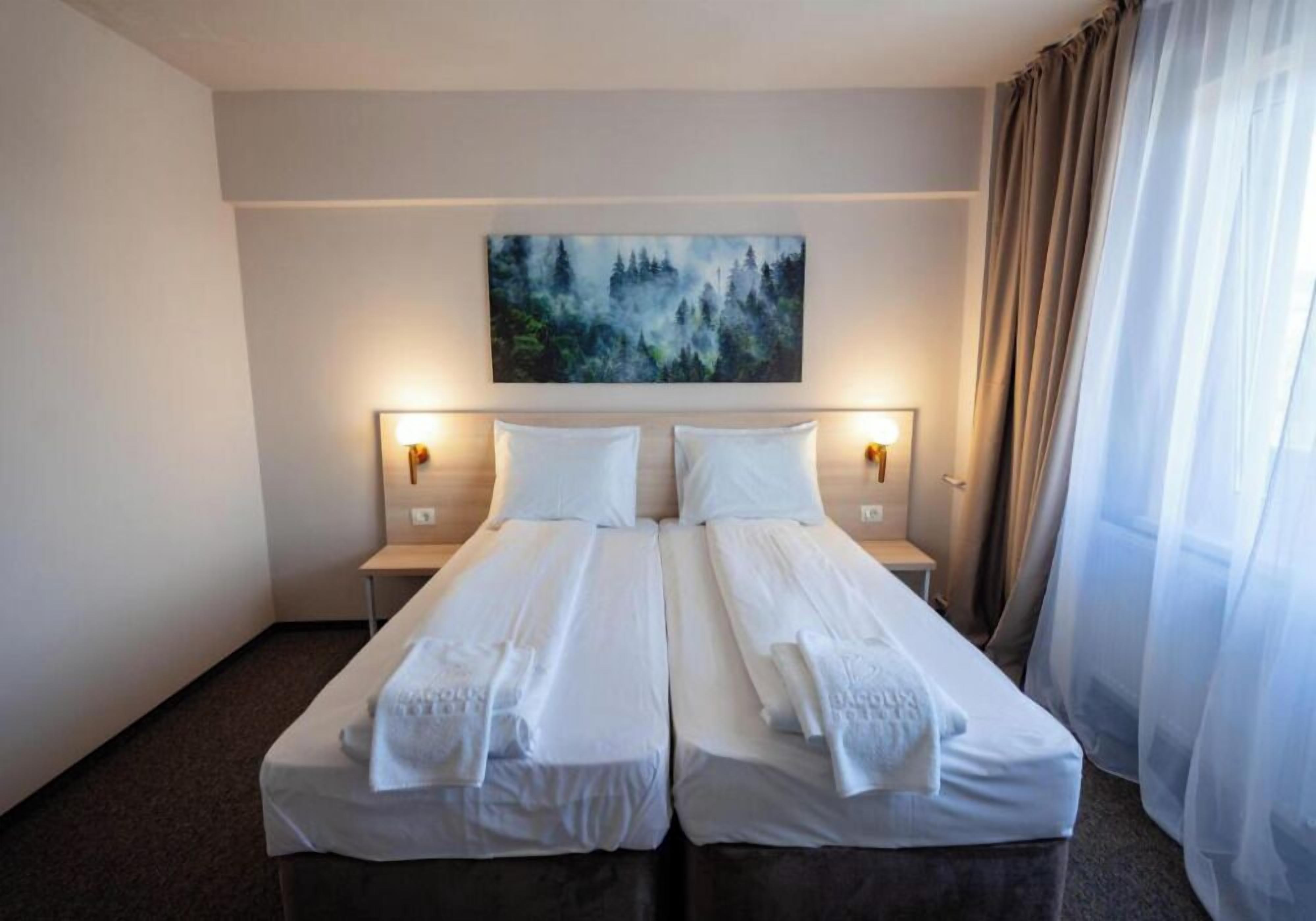 A double bed with white linens, two bedside tables with lamps, a painting on the wall, and sheer curtains on the window.
