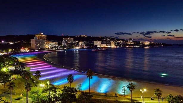 A coastal cityscape at night with a brightly lit beach and a city skyline in the background.