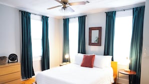 Quarto de hotel com cama, cômoda, mesa de cabeceira com abajur, ventilador de teto e janela com cortinas.
