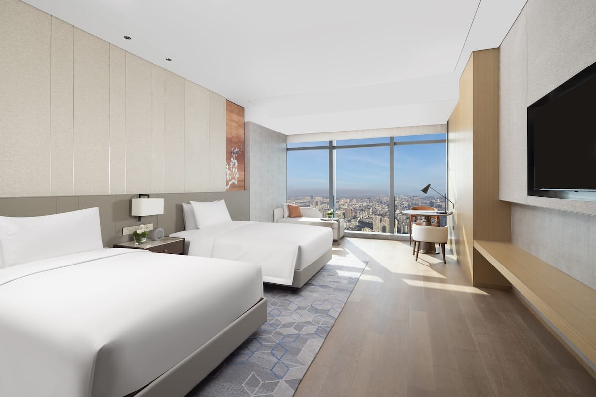 A modern hotel room with two beds, a desk, a chair, a TV, and a large window with a city view.
