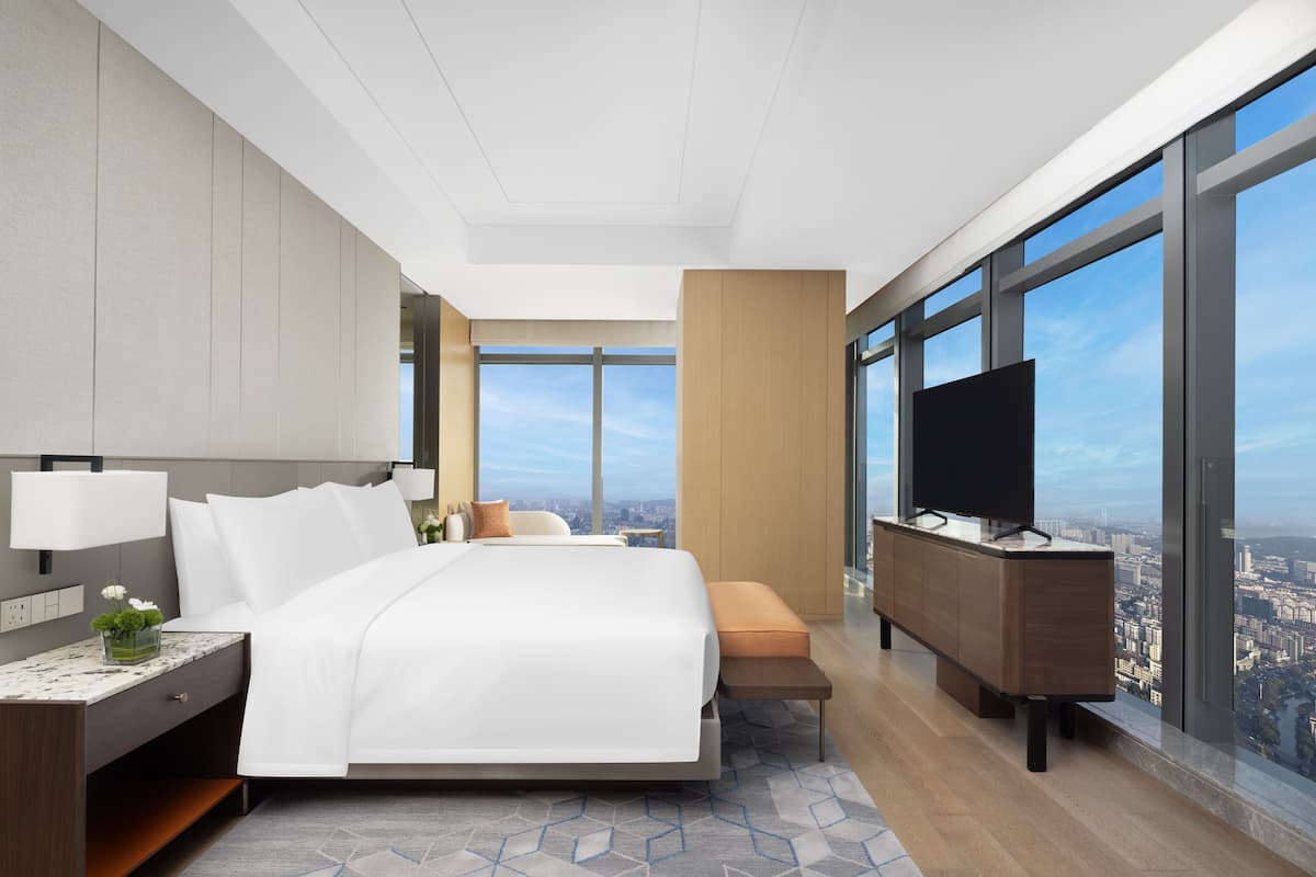 A modern hotel room with a large bed, a TV, a wooden cabinet, and a city view through large windows.