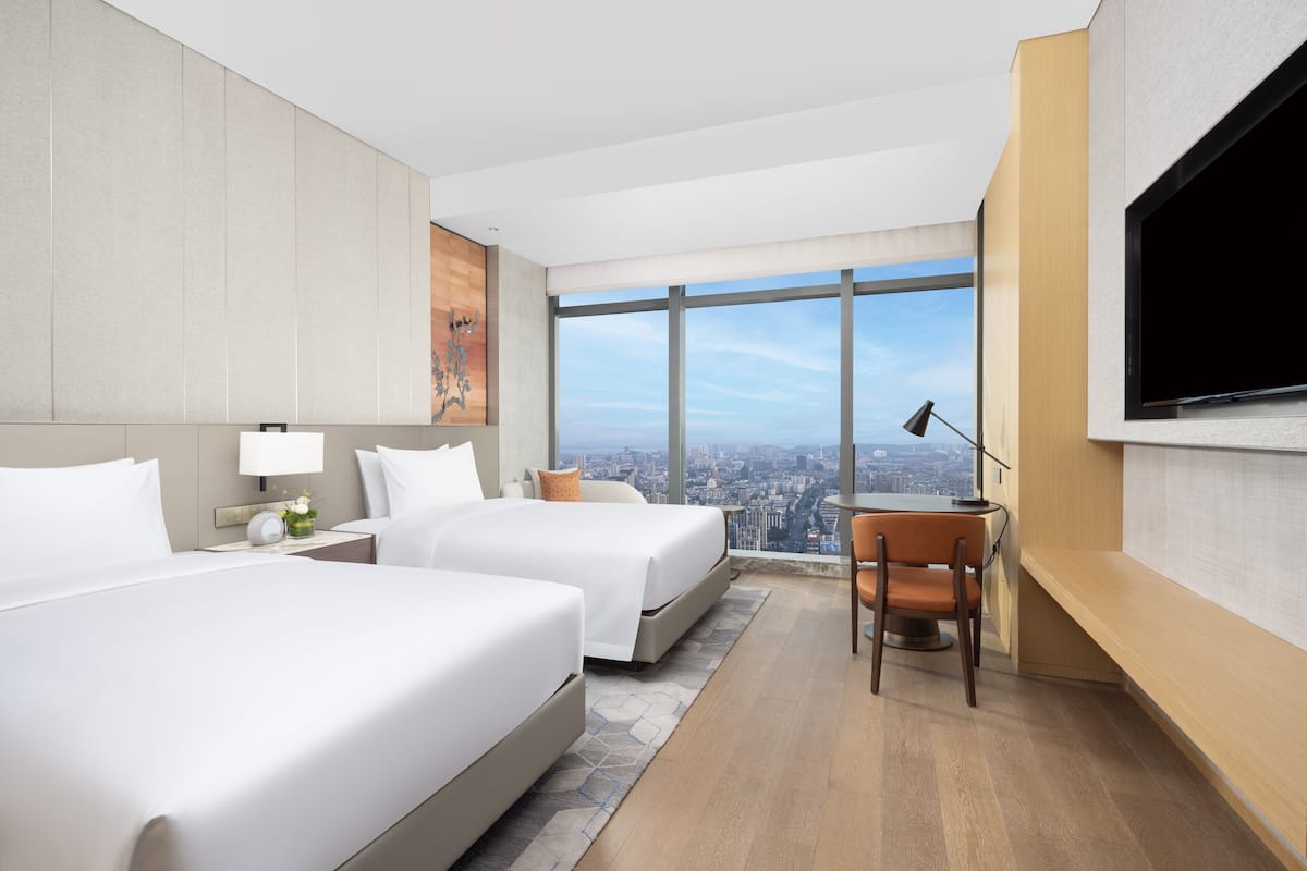 A modern hotel room with two beds, a desk, a chair, and a large TV.