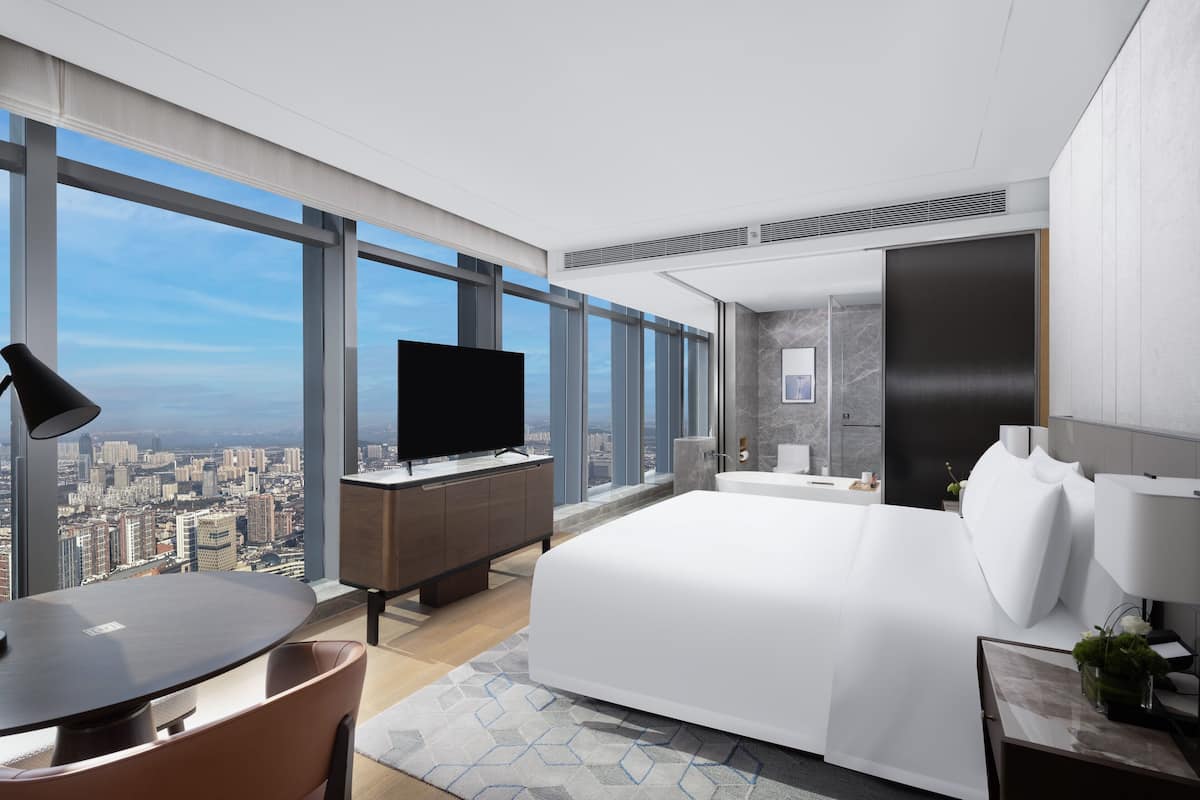 A modern hotel room with a large bed, a desk with a flat-screen TV, a dining area, and a bathroom.