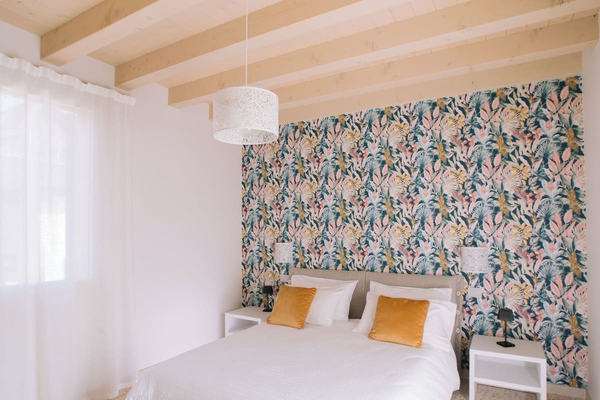 Une chambre avec un grand lit, des tables de chevet blanches, un papier peint à motifs et un plafond en bois.