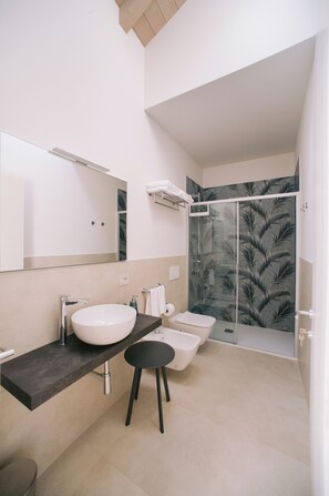 Une salle de bain moderne avec un grand miroir, un lavabo blanc, un bidet et une douche dotée d’une porte en verre ornée d’un motif de feuilles.