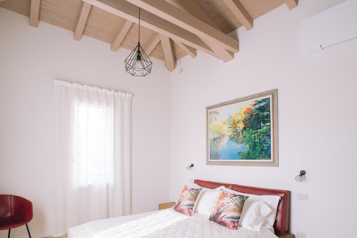 Une chambre avec un plafond en bois, un lit avec une literie blanche, une chaise rouge et un tableau accroché au mur.