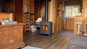 A cozy cabin interior with a wood-burning stove, wooden furniture, and a view through a window.