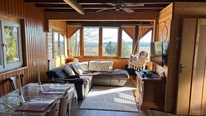 A cozy cabin interior with a wooden dining table, a sofa, a large window with a view of the outdoors, and a ceiling fan.