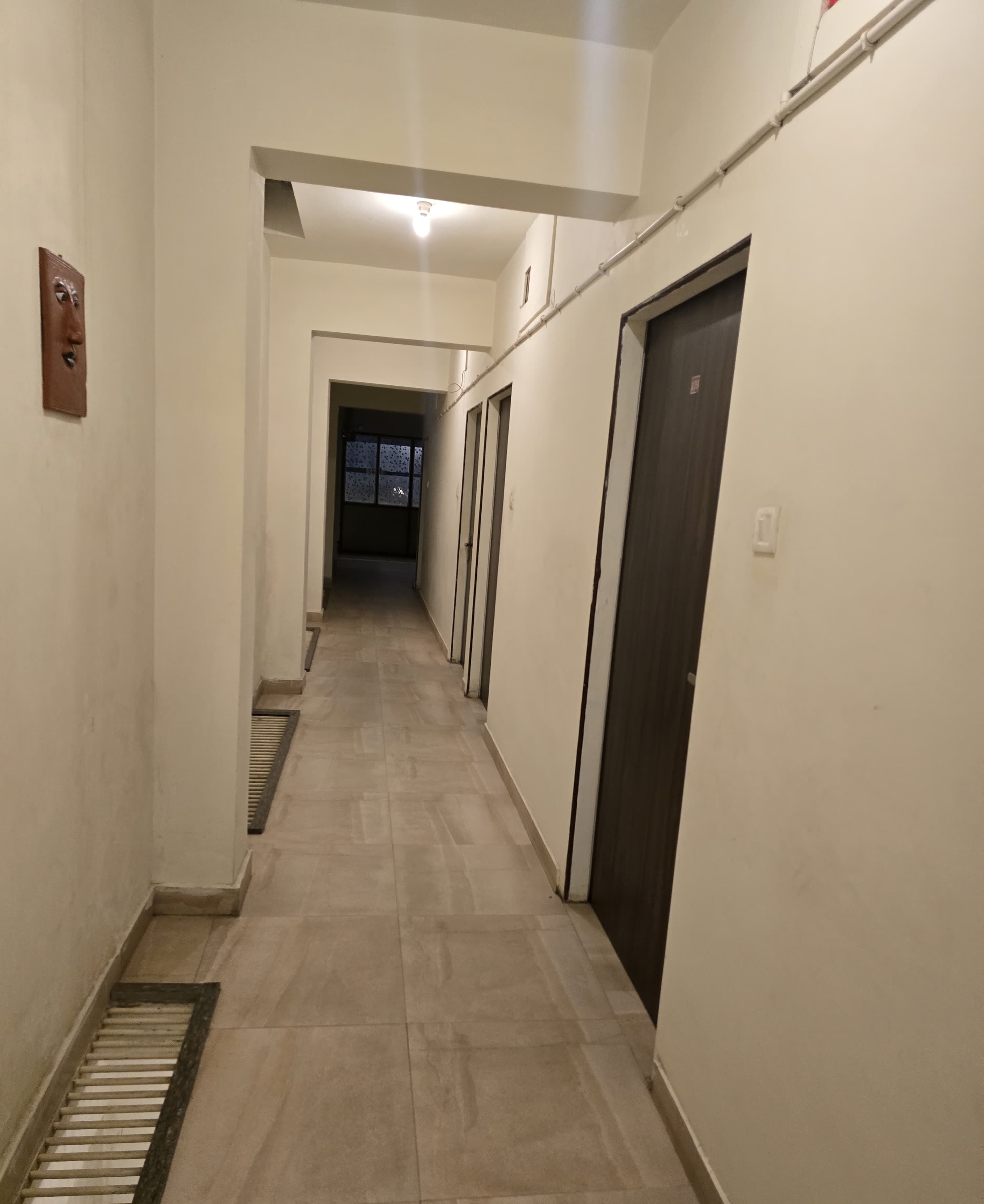 Un couloir étroit et bien éclairé, avec un sol carrelé, une grille de ventilation sur le côté gauche et une porte sur le côté droit.