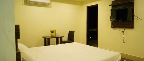 A hotel room with a bed, a small table with a plant, a chair, an air conditioning unit, and a TV mounted on the wall.