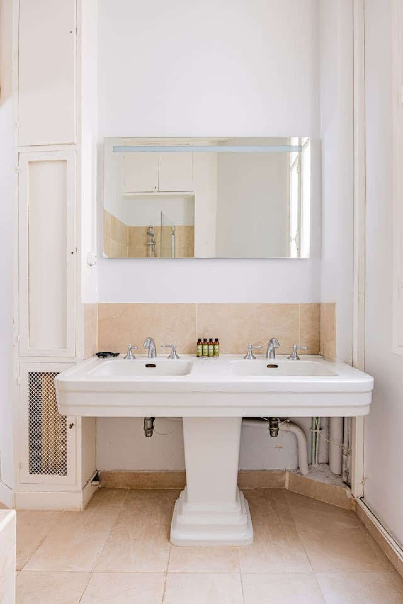 A bathroom with a white pedestal sink, a large mirror, and beige tiled walls.