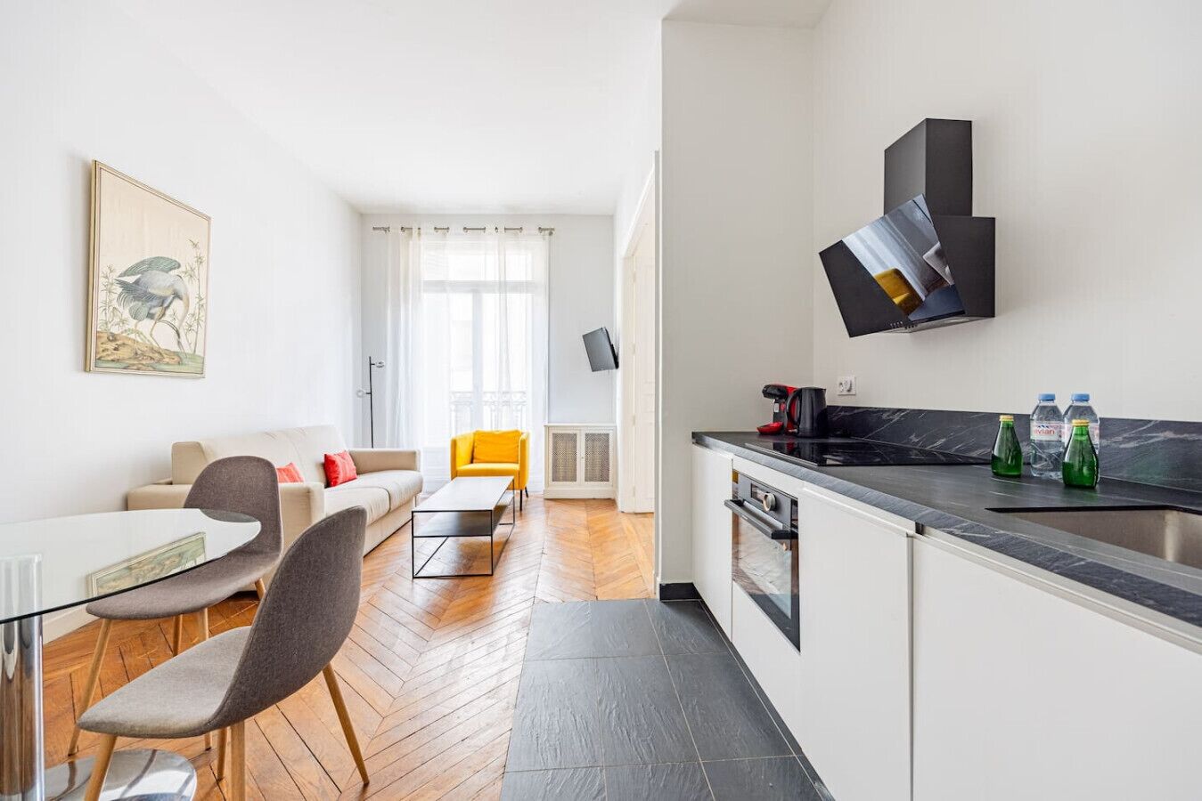 A modern apartment with a dining area, a living room with a sofa and a yellow chair, a kitchen with a black countertop and a stove, and a large window with sheer curtains.