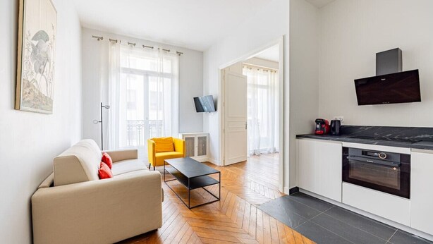 A modern living room with a sofa, a yellow chair, a coffee machine, and a TV on the wall.
