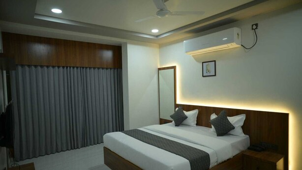 A modern hotel room with a large bed, a ceiling fan, an air conditioning unit, and a window with curtains. - Hotel Aagman (Palanpur)
