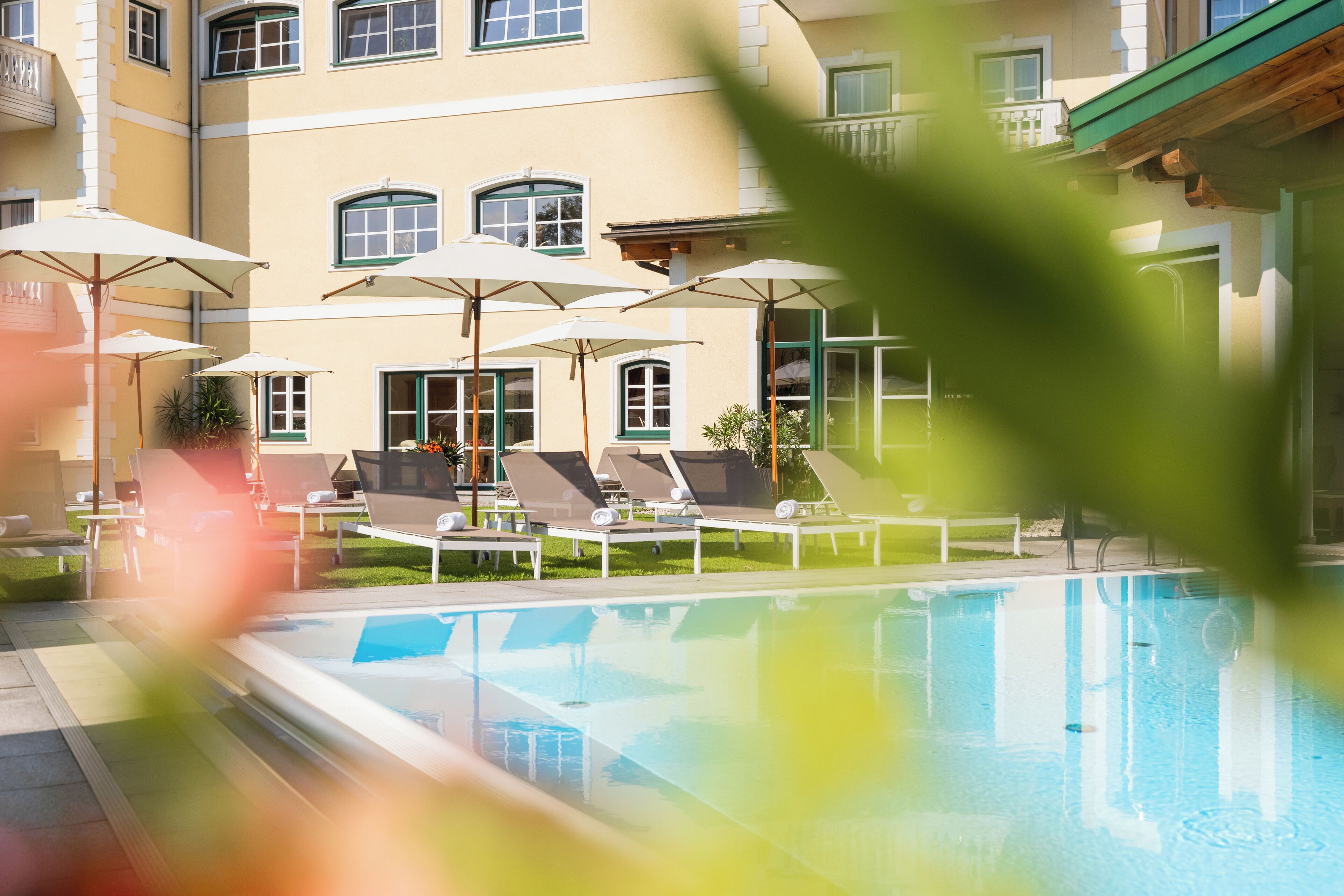 A pool area with lounge chairs and umbrellas, a building with multiple windows and balconies, and a clear blue pool.