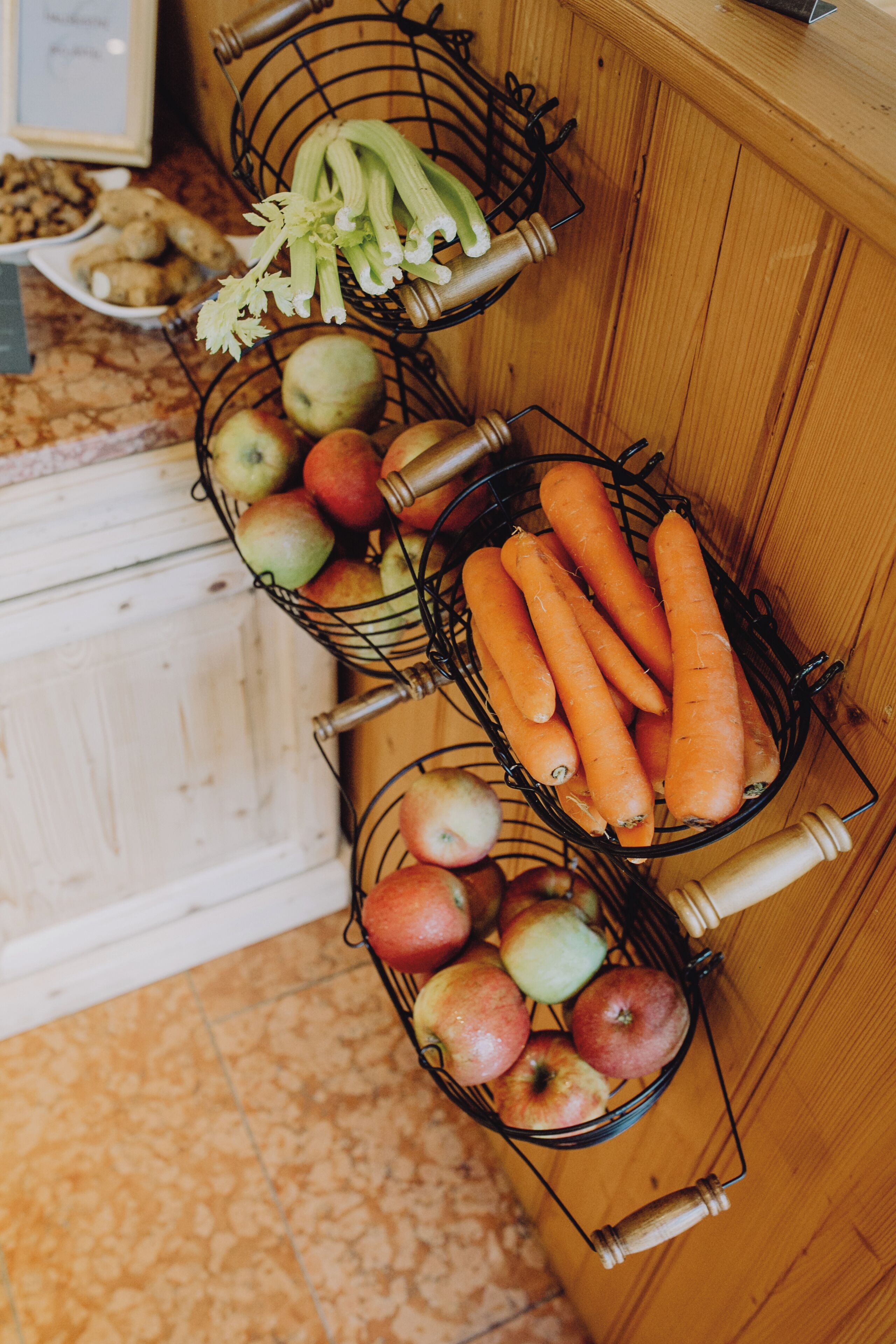 Three wire baskets containing apples, carrots, and celery on a wooden cabinet.