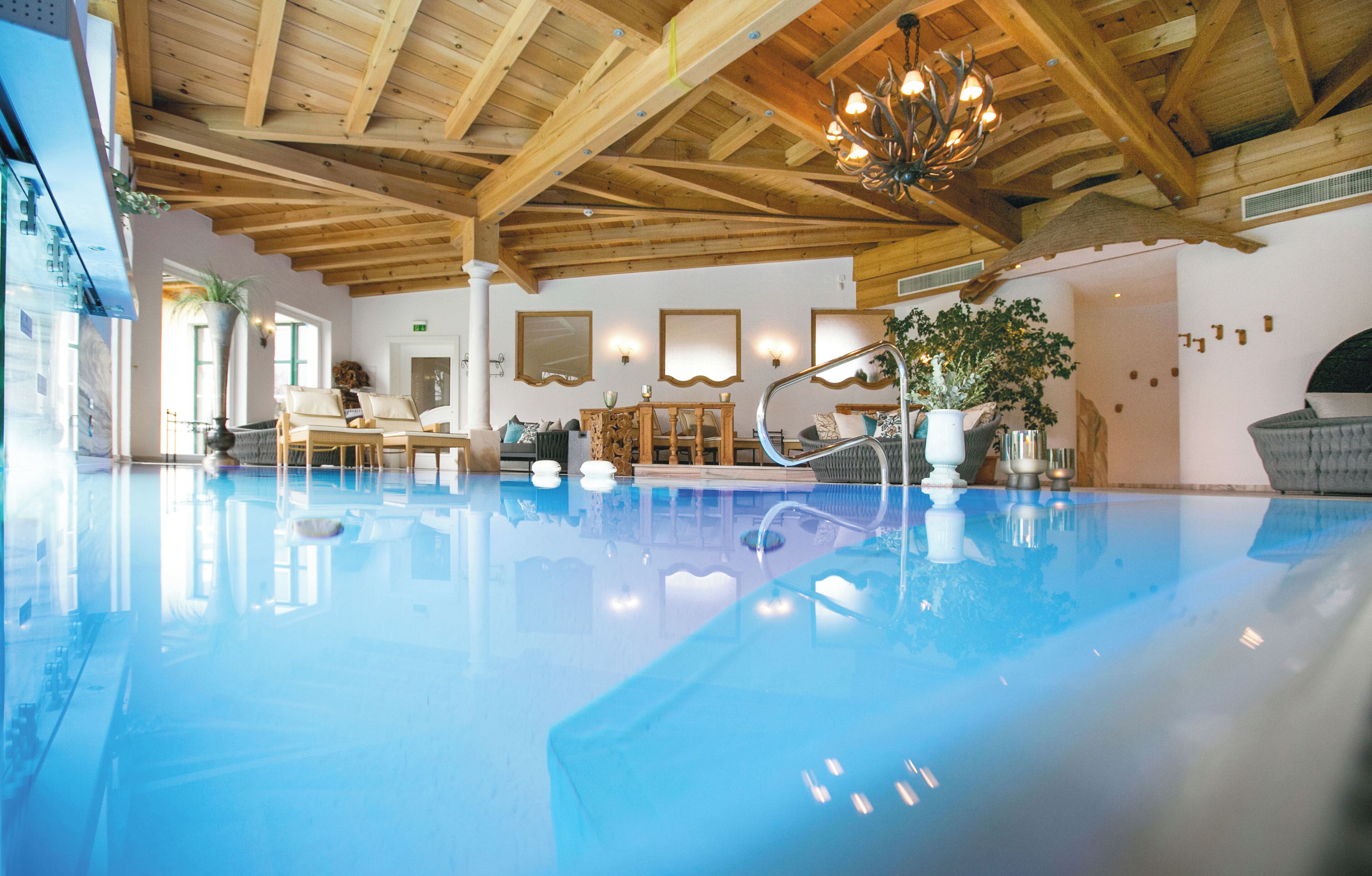 A spacious indoor swimming pool with wooden beams and a chandelier, surrounded by seating and potted plants.
