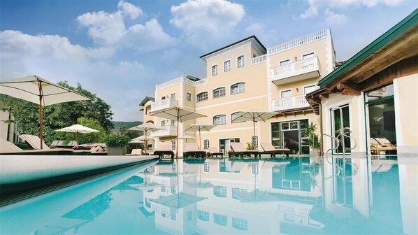A pool area with lounge chairs and umbrellas, surrounded by multi-story buildings with balconies. - Hotel Eichingerbauer (Sankt Lorenz)