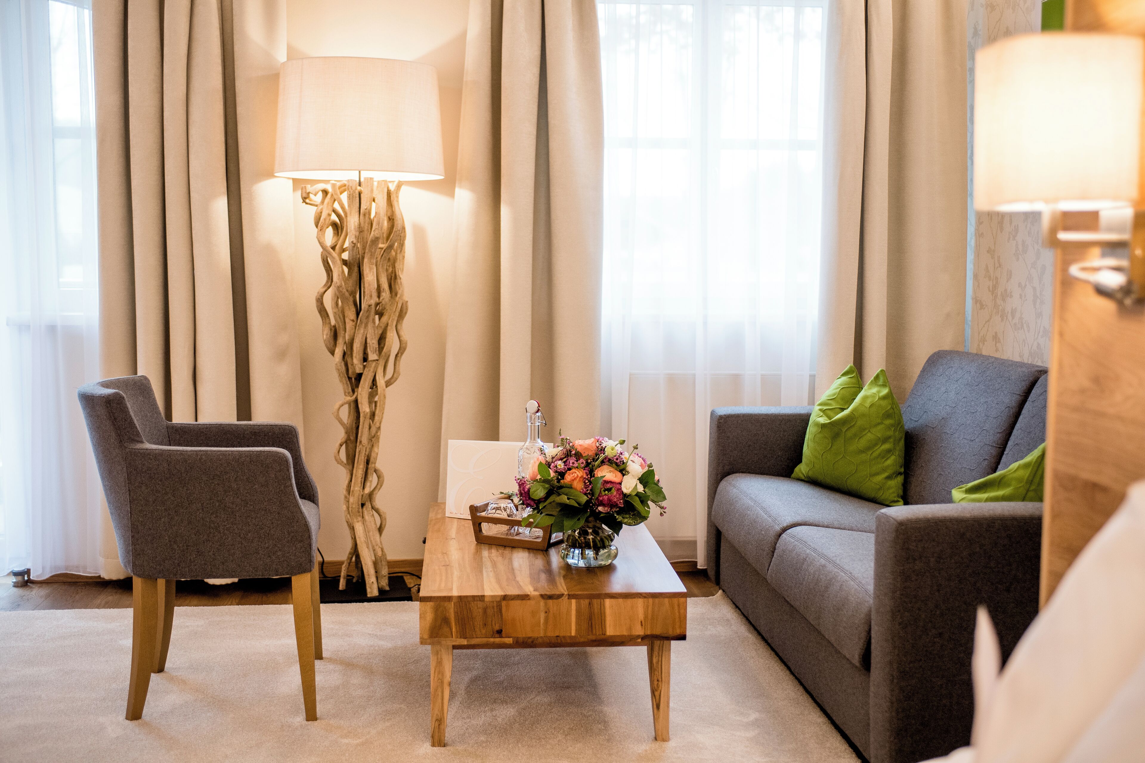 En stue med et træbord, to grå lænestole, en grå sofa med grønne puder, en gulvlampe og en blomstervase.