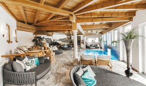 A spacious indoor area with a wooden ceiling, a stone floor, and a pool. There are seating areas with cushions and a fireplace. - Hotel Eichingerbauer (Sankt Lorenz)
