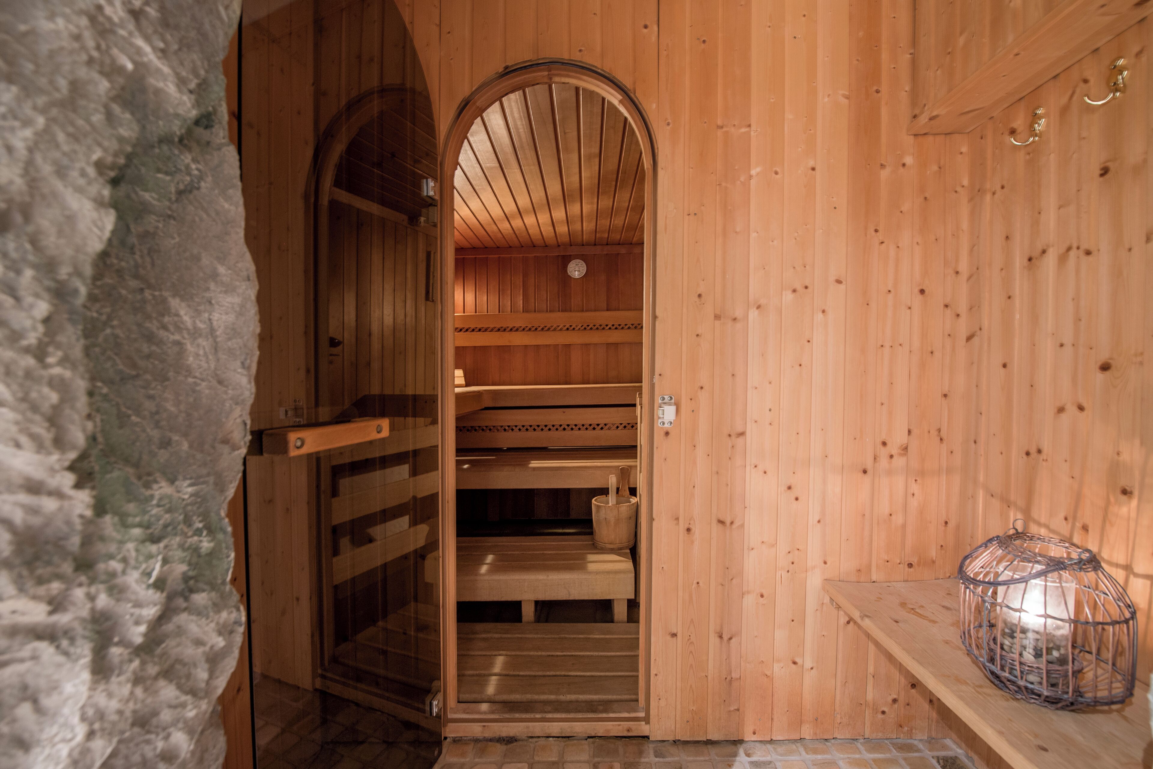 A sauna with wooden walls and benches, a stone wall on one side, and a metal container on a shelf.