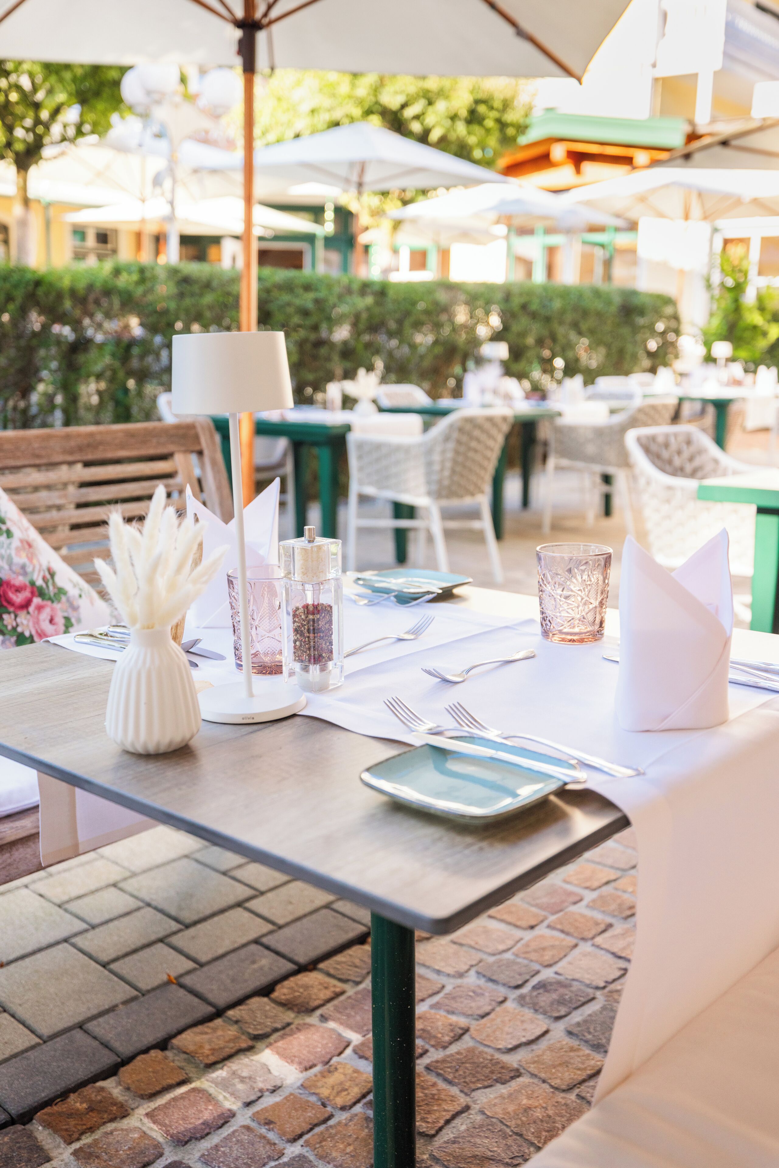 An outdoor dining area with tables set for two, featuring white tablecloths, neatly arranged cutlery, and glassware.