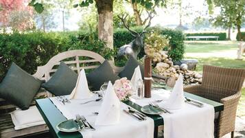 An outdoor dining setup with a table set for two, surrounded by greenery and a view of a lake.