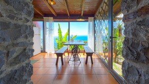 A stone-walled room with a wooden ceiling, a table with benches, and a view of the ocean through large windows. - Cottage Sapateiro, a shelter in the nature! (Arco da Calheta)