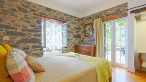 A bedroom with a stone wall, a bed with a yellow blanket, a dresser, and a view of the outdoors through a glass door. - Cottage Sapateiro, a shelter in the nature! (Arco da Calheta)