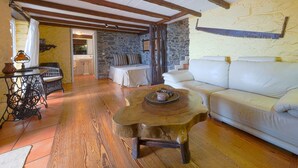 A cozy living room with a stone wall, a wooden ceiling, a white sofa, a wicker table, and a wooden coffee table. - Cottage Sapateiro, a shelter in the nature! (Arco da Calheta)