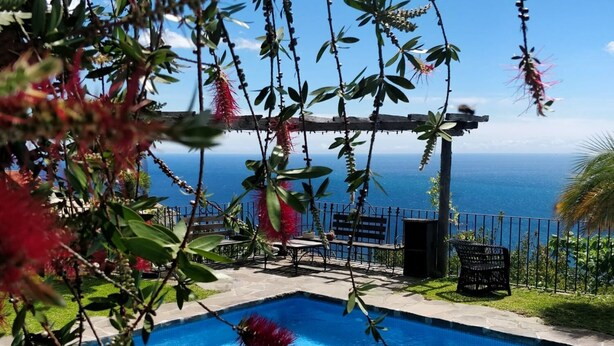 A clear blue pool with a wooden deck, a metal pergola, and a view of the ocean. - Cottage Sapateiro, a shelter in the nature! (Arco da Calheta)