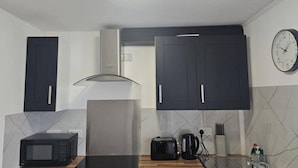 A compact kitchen with dark cabinets, a microwave, a toaster, a kettle, and a clock on the wall.