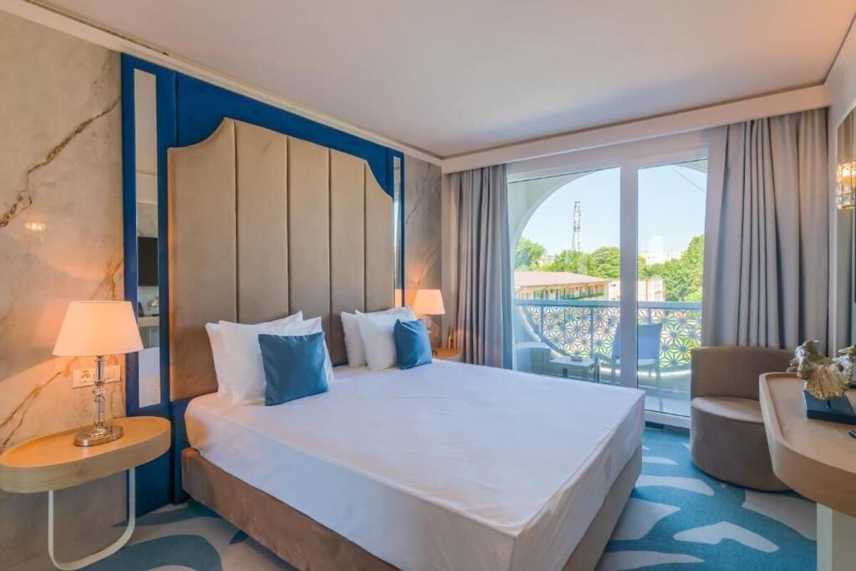 Habitación de hotel con una cama grande, dos mesitas de noche con lámparas, una silla y un balcón con vistas.
