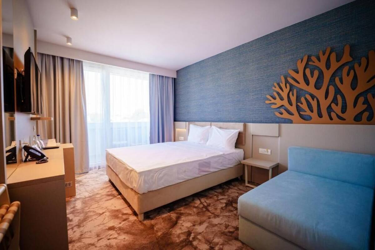 Habitación de hotel con cama, escritorio con teléfono, silla, televisor y una ventana grande con cortinas.