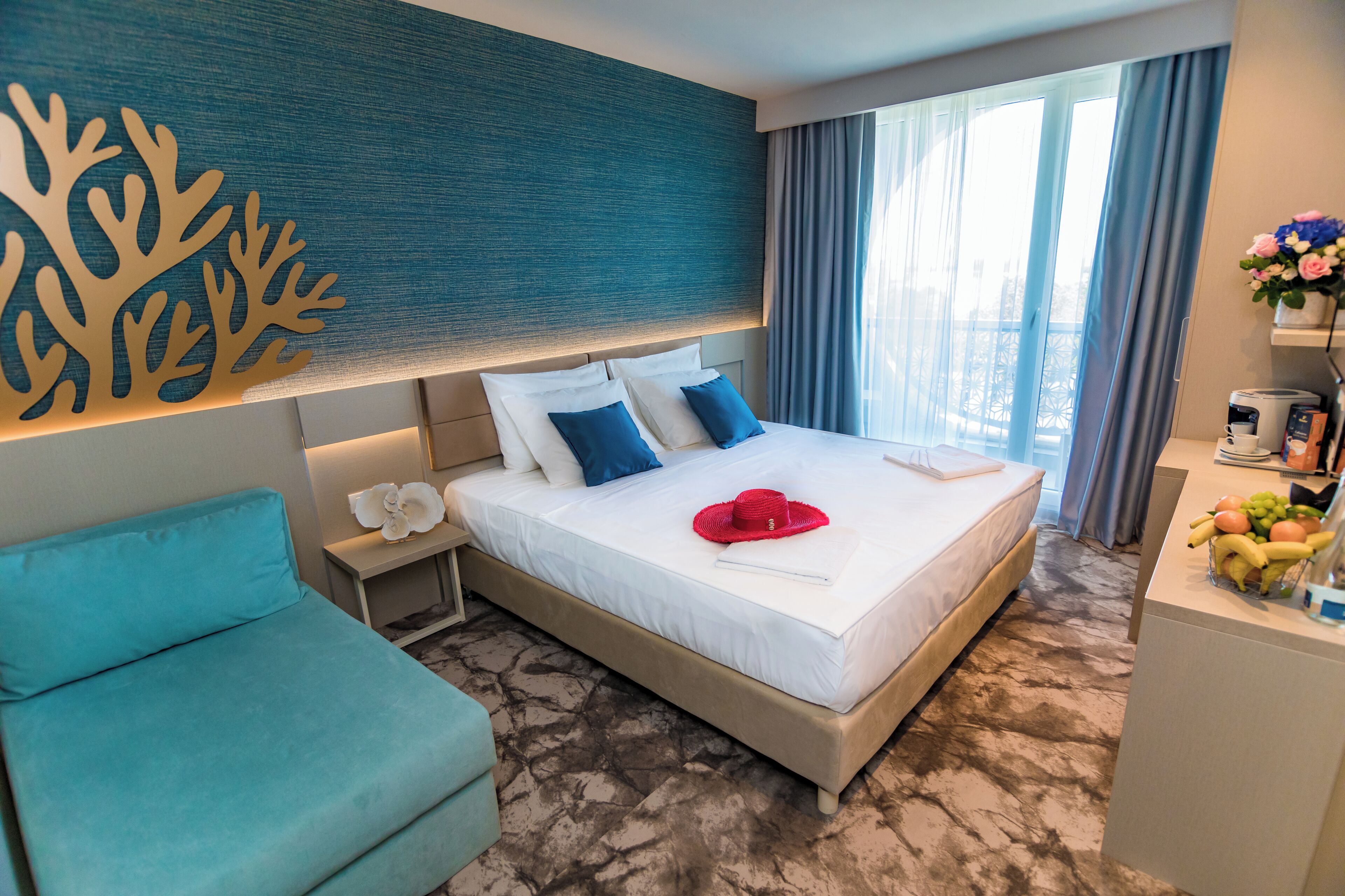 A hotel room with a bed, a desk with a fruit bowl, a chair, and a decorative wall piece.