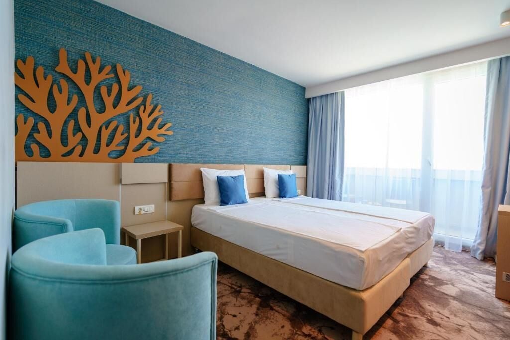 A hotel room with a bed, two blue chairs, a nightstand, a wall art piece, and a large window with curtains.