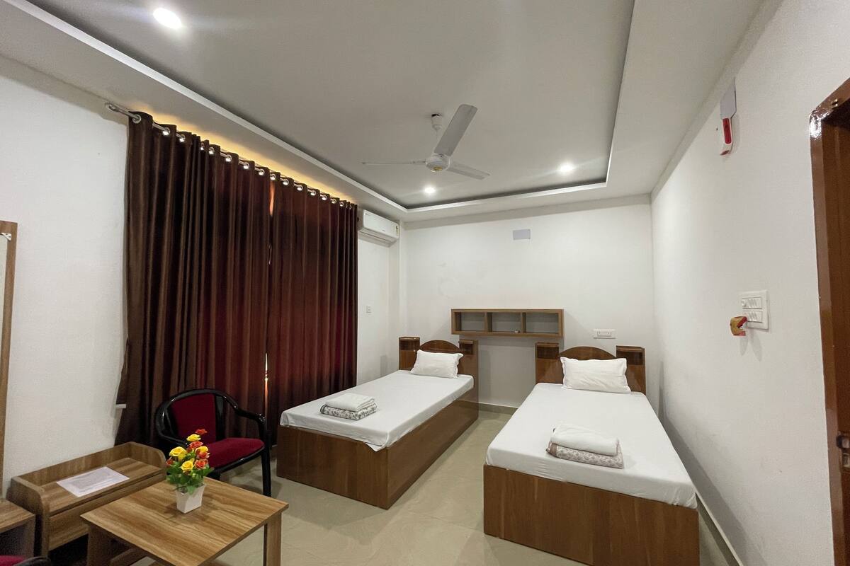 A hotel room with two single beds, a desk with a chair, a small table with a vase of flowers, and a ceiling fan.